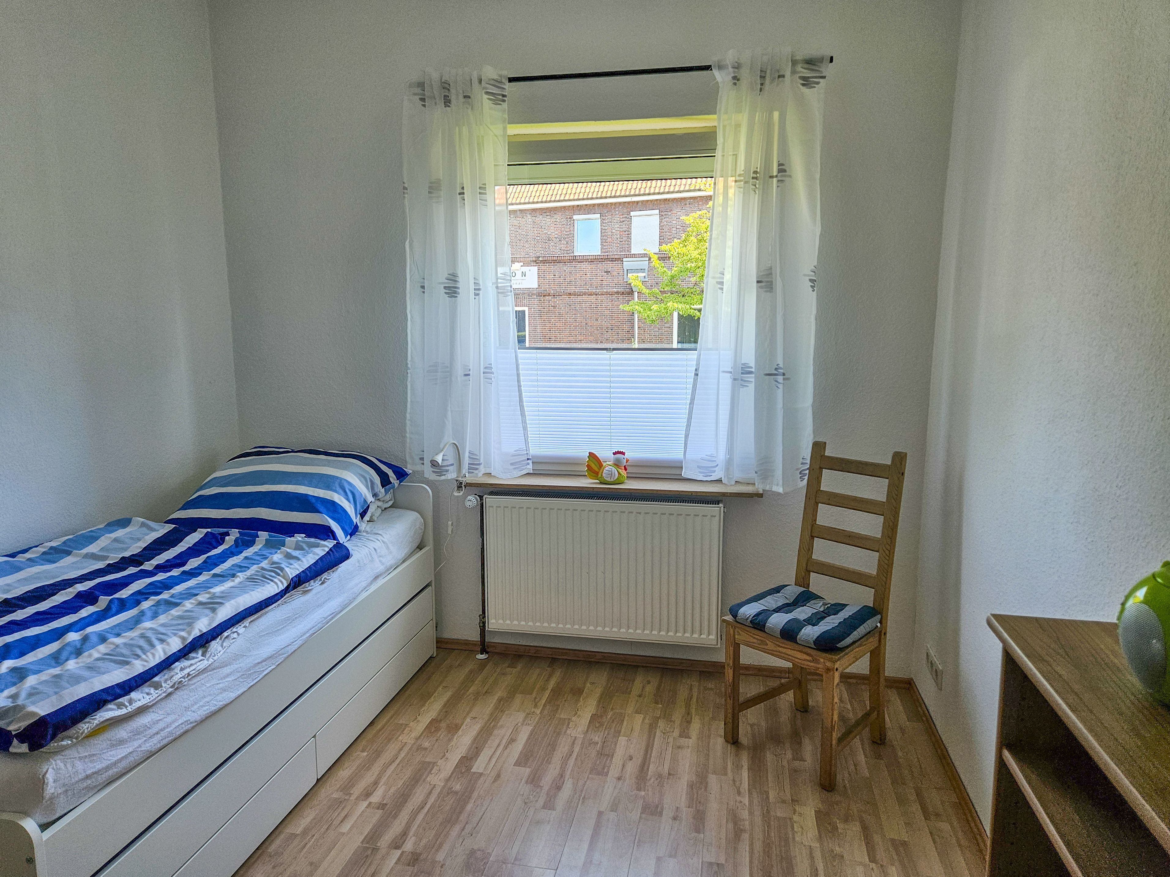 A small, single-bed room with a wooden floor, a radiator, a window with sheer curtains, a wooden chair with a cushion, and a small table with a vase.