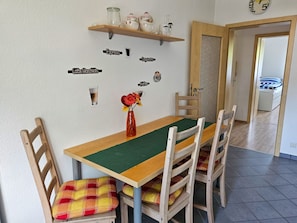 Uma área de refeições com mesa de madeira, almofadas xadrez e um vaso com flores. Há etiquetas fixadas na parede para diferentes tipos de café.