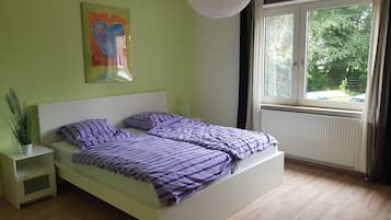 Un dormitorio con cama, mesita de noche, planta en maceta, ventana con cortinas y lámpara colgante.