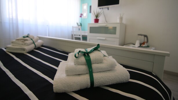 A neatly made bed with folded towels and a green ribbon, a white bedside table with a potted plant, and a TV mounted on the wall. - Sette sorelle b&b (Cosenza)