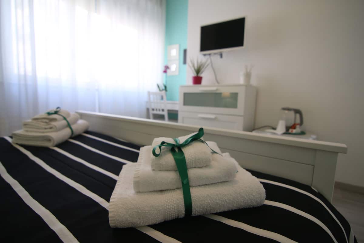 A neatly made bed with folded towels and a green ribbon, a white bedside table with a potted plant, and a TV mounted on the wall.