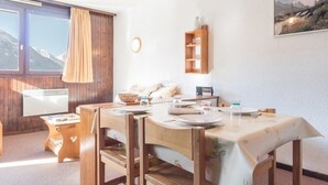 Un coin repas avec une table en bois dressée pour deux personnes, une petite table d’appoint et une vue sur les montagnes par la fenêtre.