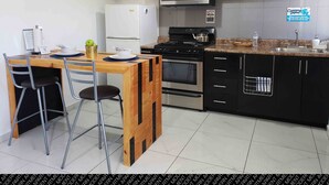 Une cuisine moderne avec des meubles noirs, un four à micro-ondes en acier inoxydable, un réfrigérateur blanc, une table à manger en bois avec deux chaises et un sol carrelé.