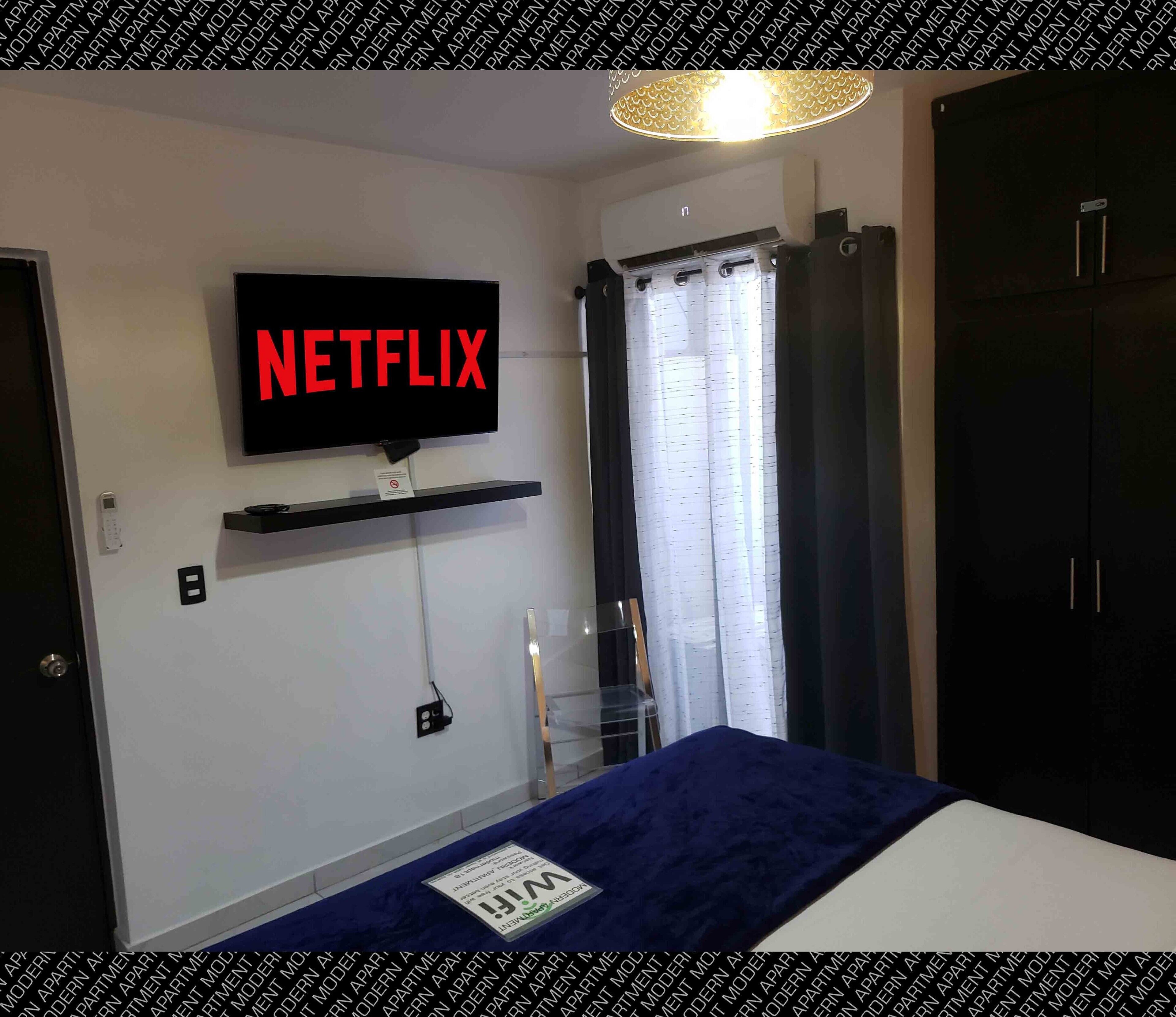 A bedroom with a mounted TV displaying "NETFLIX," a bedside table, a bed with a blue cover, a wardrobe, and a window with curtains.