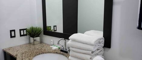 A bathroom with a granite countertop, a sink, a mirror, a plant, and a stack of towels.