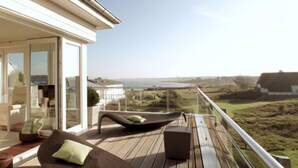 Een houten terras met een lounger en kussens, uitkijkend op een kustlandschap.