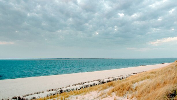 Een strand met een vuurtoren op een grasheuvel, een zandstrand en een bewolkte lucht.