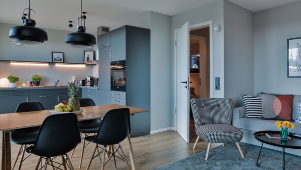 A modern kitchen with a dining area, a living room with a sofa and a coffee table, and a hallway leading to a bedroom. - Bett & Bude Boardinghouse (Sylt)