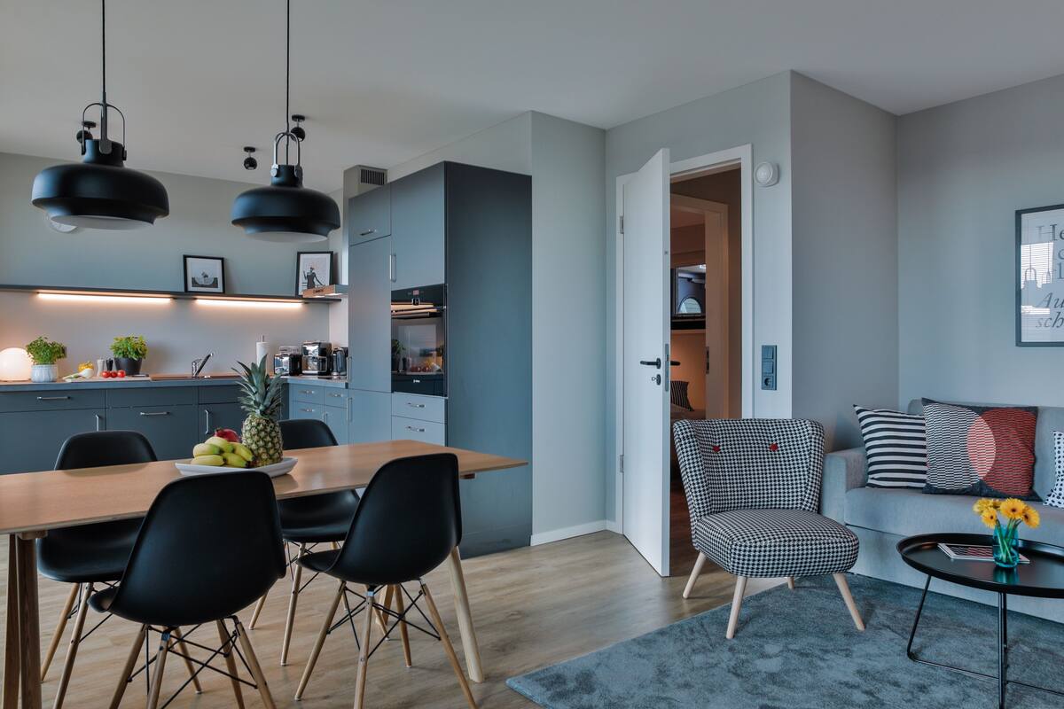 A modern kitchen with a dining area, a living room with a sofa and a coffee table, and a hallway leading to a bedroom.