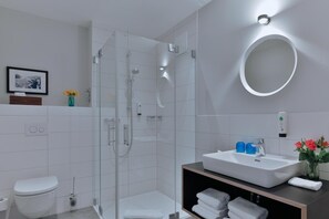 A modern bathroom with a glass-enclosed shower, a white sink with blue soap dispensers, and a round mirror on the wall. - Bett & Bude Boardinghouse (Sylt)