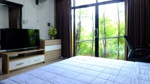 A bedroom with a bed, a TV on a wooden stand, a window with curtains, and a view of greenery outside. - Sirimongkol Resort (Surat Thani)
