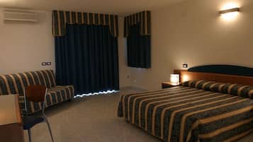 A hotel room with a bed, a sofa, a chair, a bedside table with a lamp, and a window with curtains.
