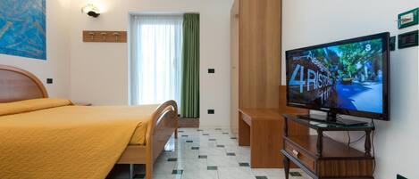 A hotel room with a bed, a television on a wooden stand, a window with curtains, and a wall-mounted light.