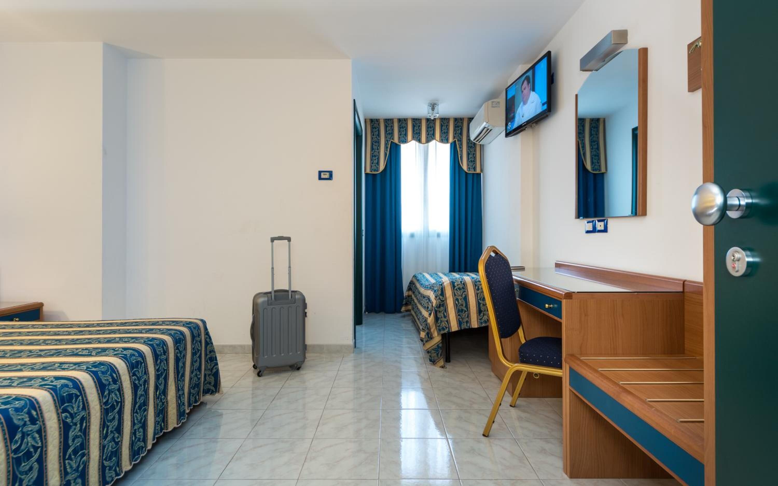 Ein Hotelzimmer mit zwei Betten, einem Schreibtisch, einem Stuhl, einem Koffer und einem an der Wand befestigten Fernseher.