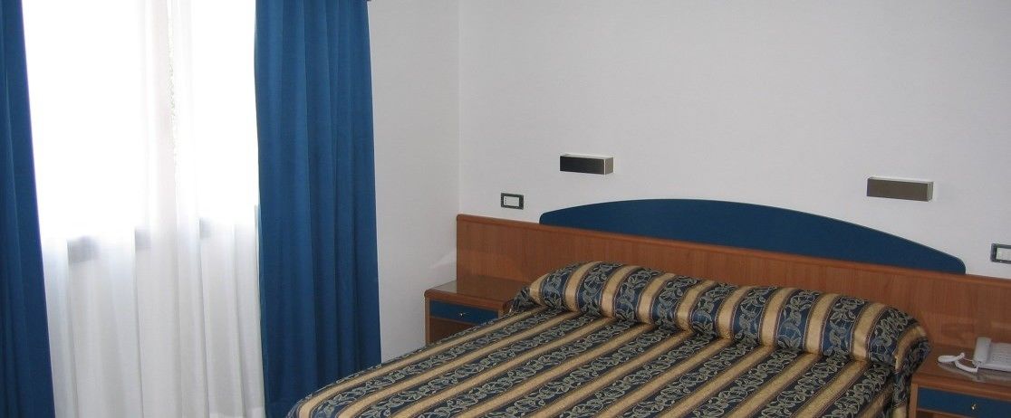Een hotelkamer met een bed, nachtkastje en een raam met gordijnen.