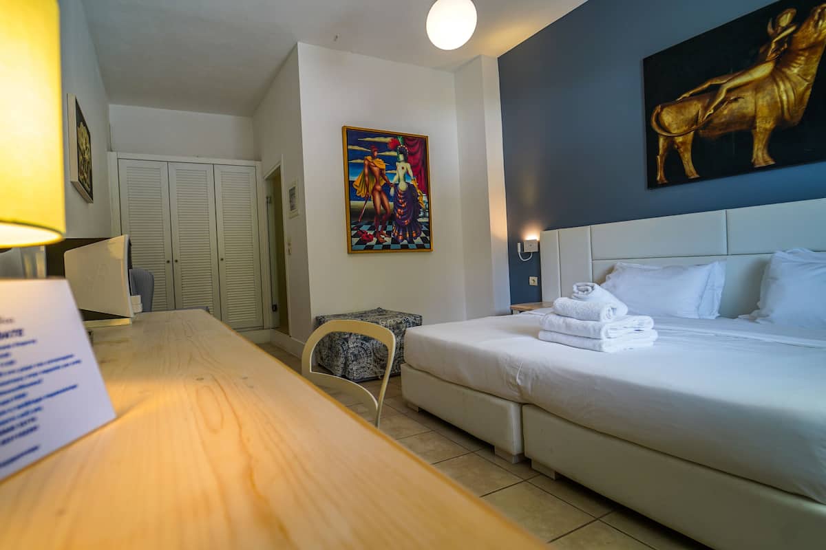 Une chambre d’hôtel avec un lit, un bureau, une chaise, une lampe, un tableau et une porte.