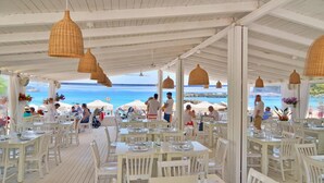 Zona pranzo all'aperto con tavoli e sedie bianche, con vista su una spiaggia con ombrelloni e persone.