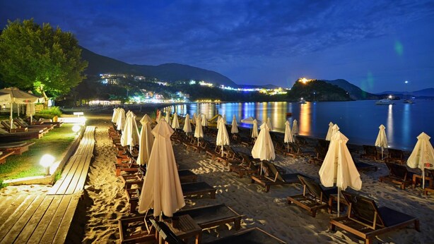 Una spiaggia con lettini e ombrelloni, un pontile di legno e una vista su una città sullo sfondo.