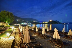 A beach with sun loungers and umbrellas, a wooden deck, and a view of a city in the background. - Cape North West (Parga)