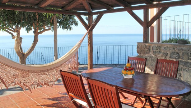 A wooden table with chairs, a hammock, and a fruit bowl on a patio overlooking the sea. - Stonecliff Cottage by Our Madeira (Calheta)