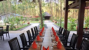 A long dining table set for a meal in an outdoor setting with chairs on both sides, surrounded by a garden and a bamboo forest.