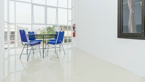 Een lichte kamer met een groot raam, een tafel met stoelen en een brandblusser aan de muur.