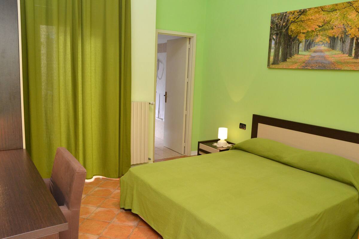 A room with a bed, a chair, a table, a nightstand with a lamp, and a painting on the wall.