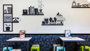A room with two tables, each with a poster and a small plant. There are colorful stools and a wall with framed pictures and a bird illustration.