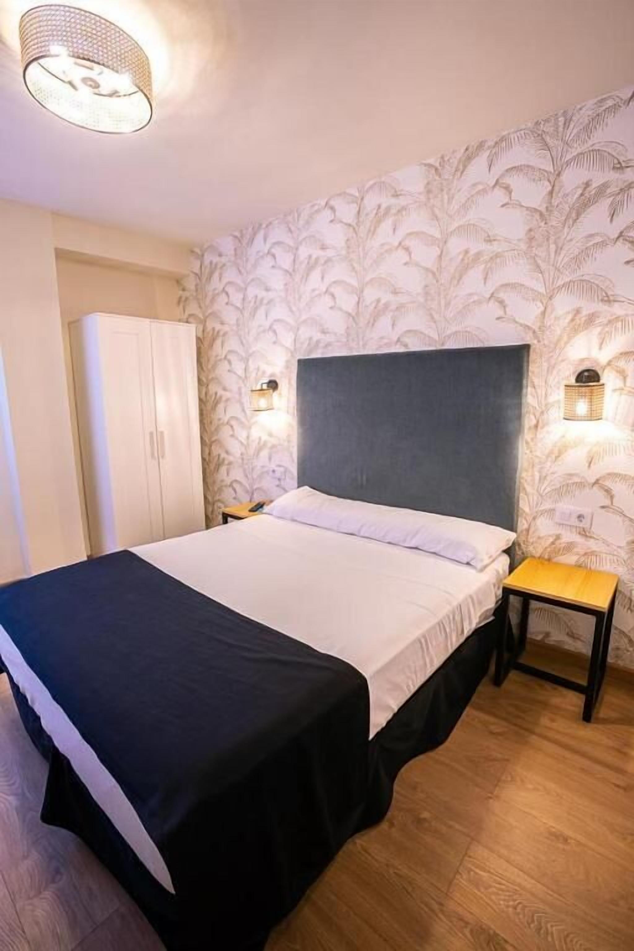 Ein Schlafzimmer mit Bett, Nachttisch, Lampe, Kleiderschrank und Wandtapete mit Muster.