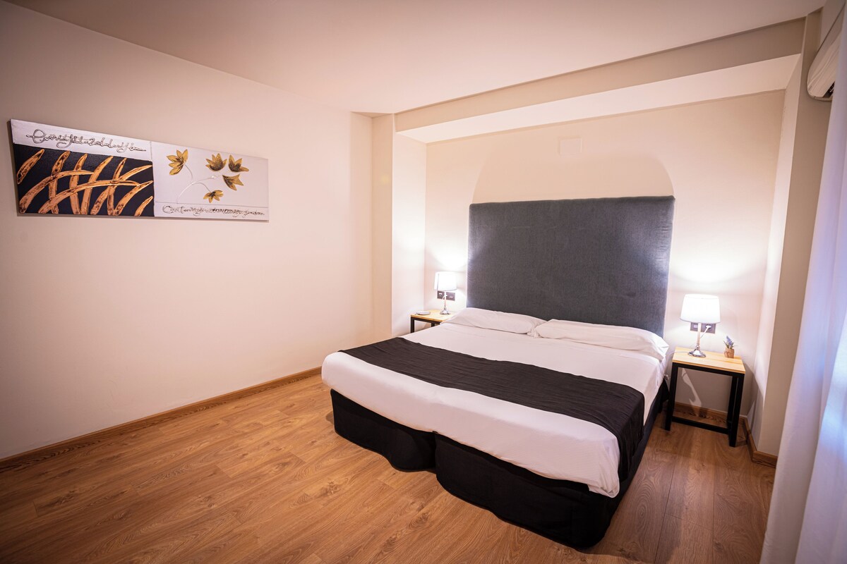 A hotel room with a bed, two bedside tables, a nightstand, a wall-mounted artwork, and a wooden floor.