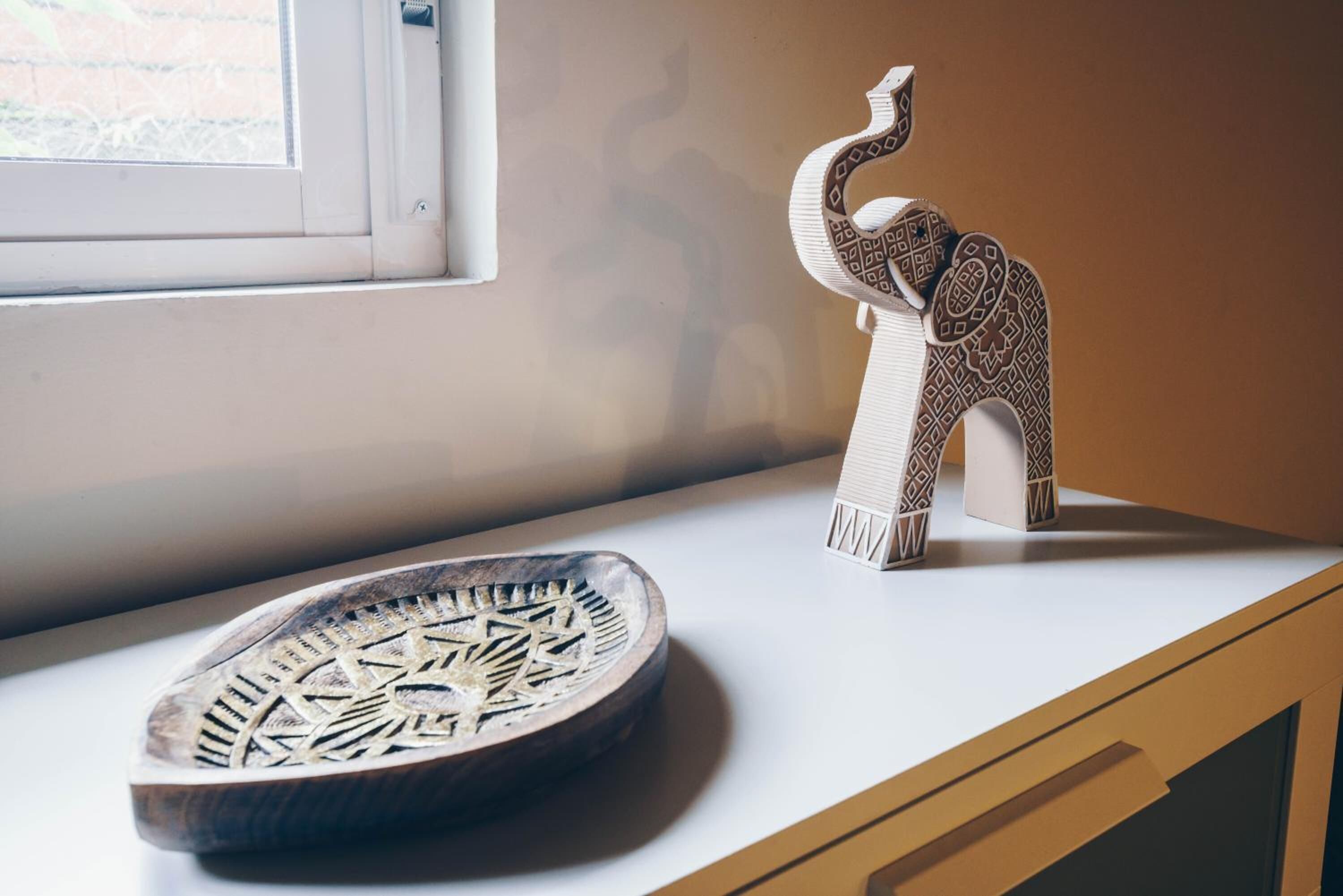 Una scultura di elefante in legno e una ciotola decorativa su una superficie bianca vicino a una finestra.