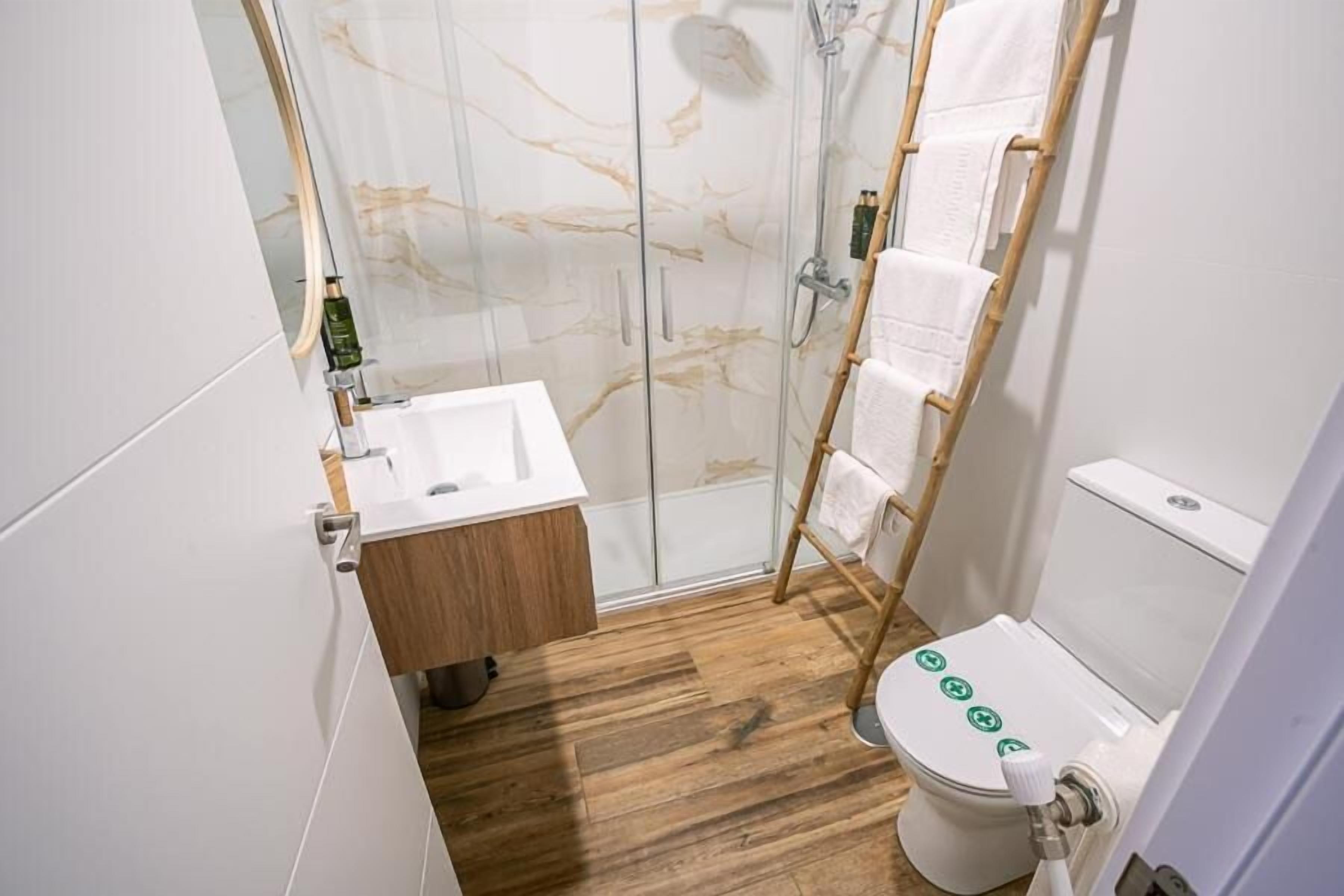 A bathroom with a glass shower enclosure, a wooden floor, a white sink, and a toilet with a bidet attachment.