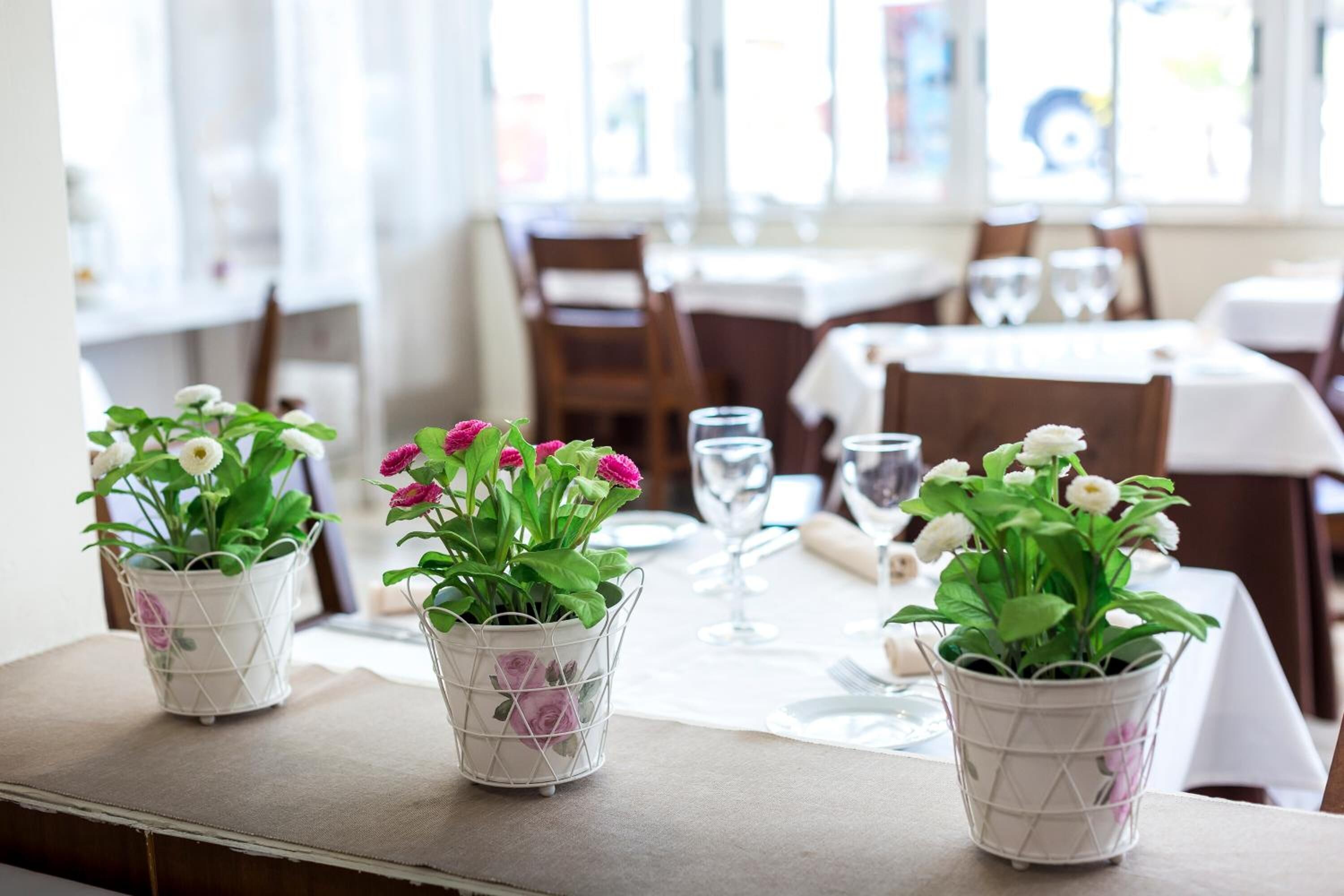 A dining area with tables set for two, each with a white tablecloth, a wine glass, and a potted plant with white and pink flowers.