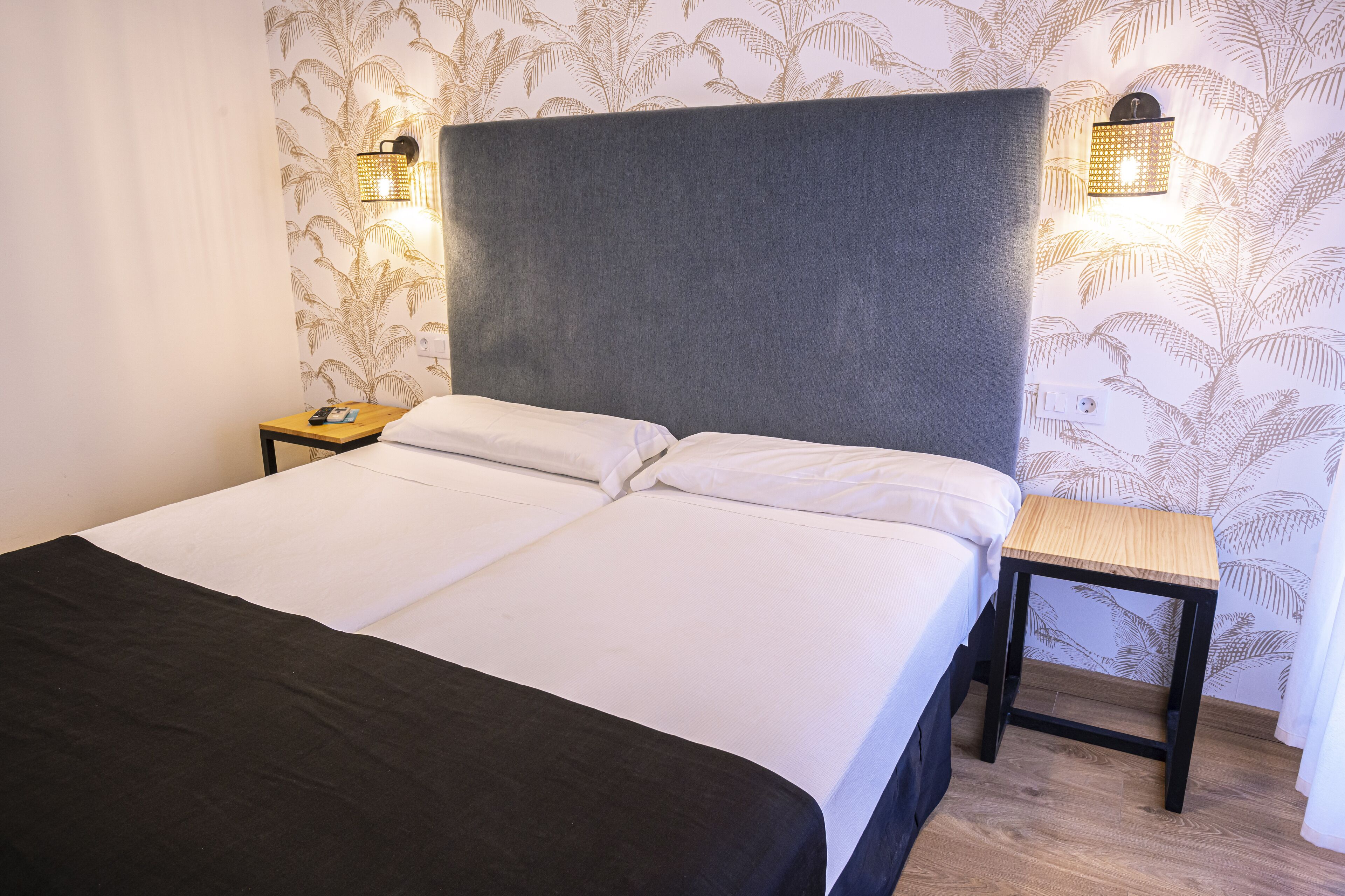 Un lit double avec une tête de lit grise, deux tables de chevet et une petite table de nuit avec une lampe.