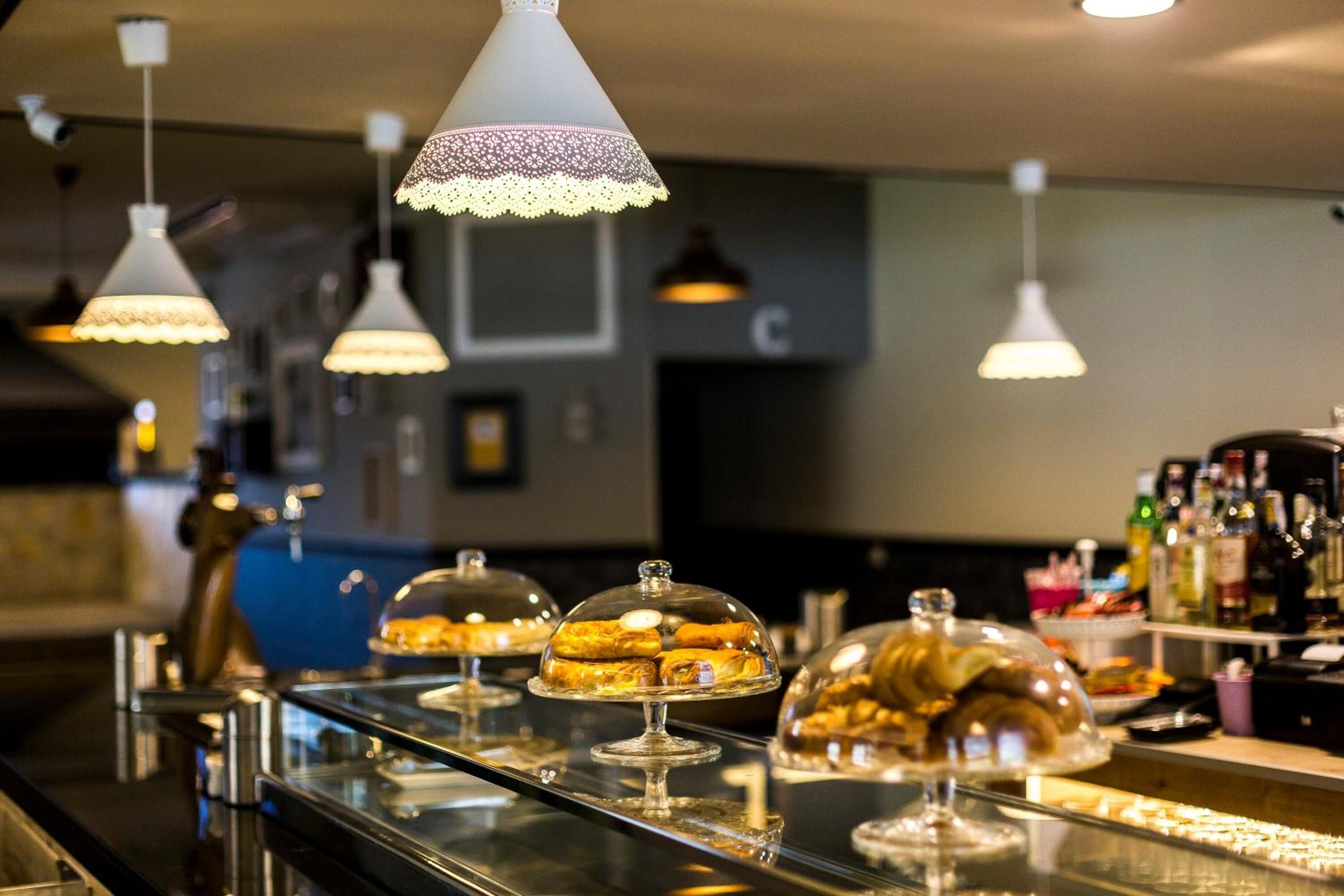 A buffet area with various pastries under glass domes, overhead pendant lights, and a bar area with bottles in the background.