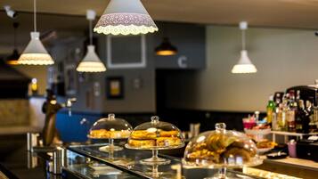 A buffet area with various pastries under glass domes, overhead pendant lights, and a bar area with bottles in the background.