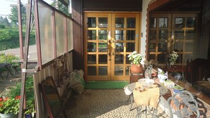 A covered porch with a wooden door, potted plants, and outdoor furniture. - Le Café Cerisier (Myoko)