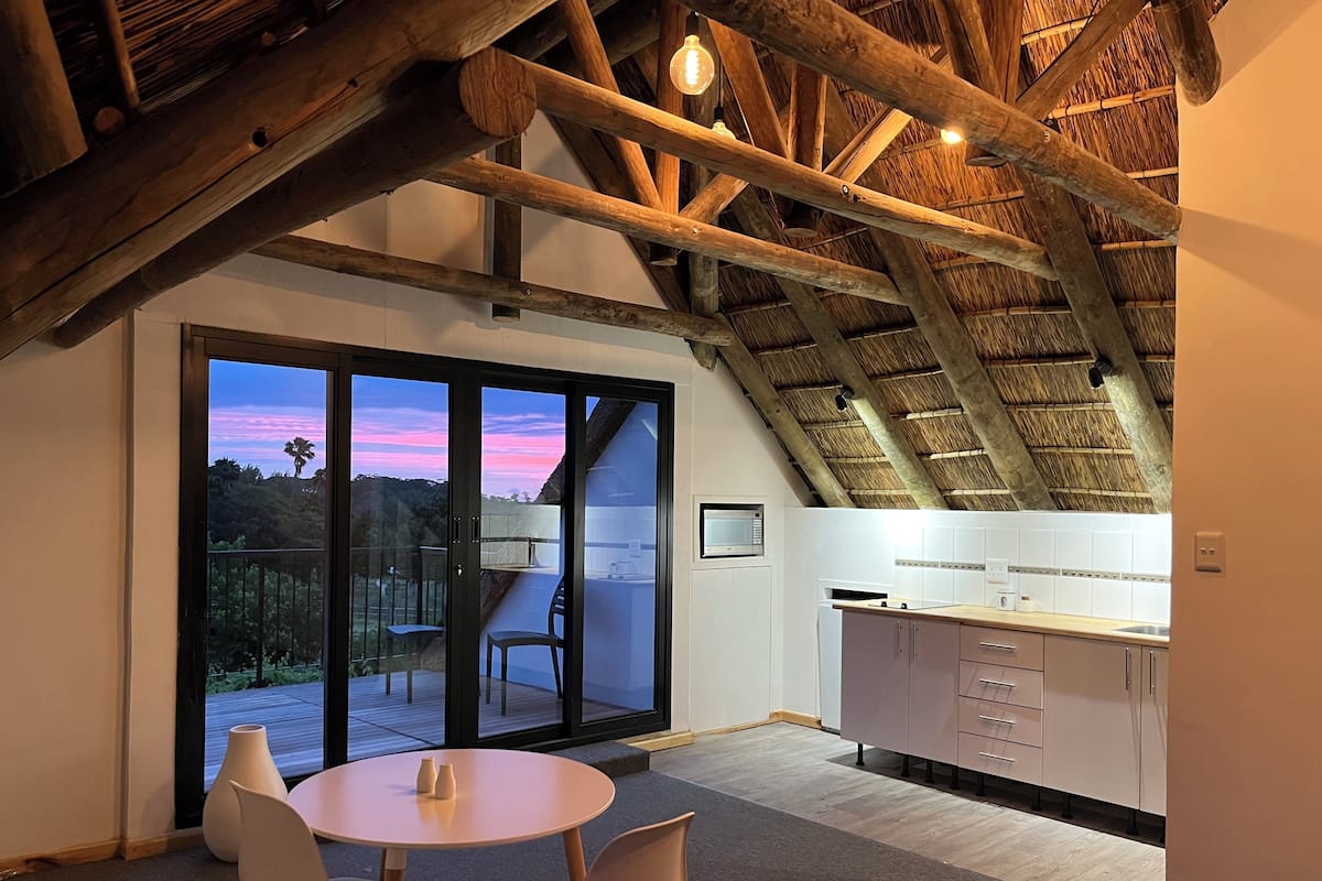 A modern kitchen with white cabinets and a wooden ceiling, a dining area with a round table and chairs, and a large sliding glass door leading to an outdoor deck.