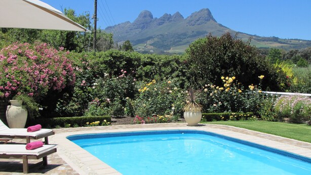 Una piscina con sillas de descanso y un gran paraguas, rodeada de jardines exuberantes y con montañas al fondo.