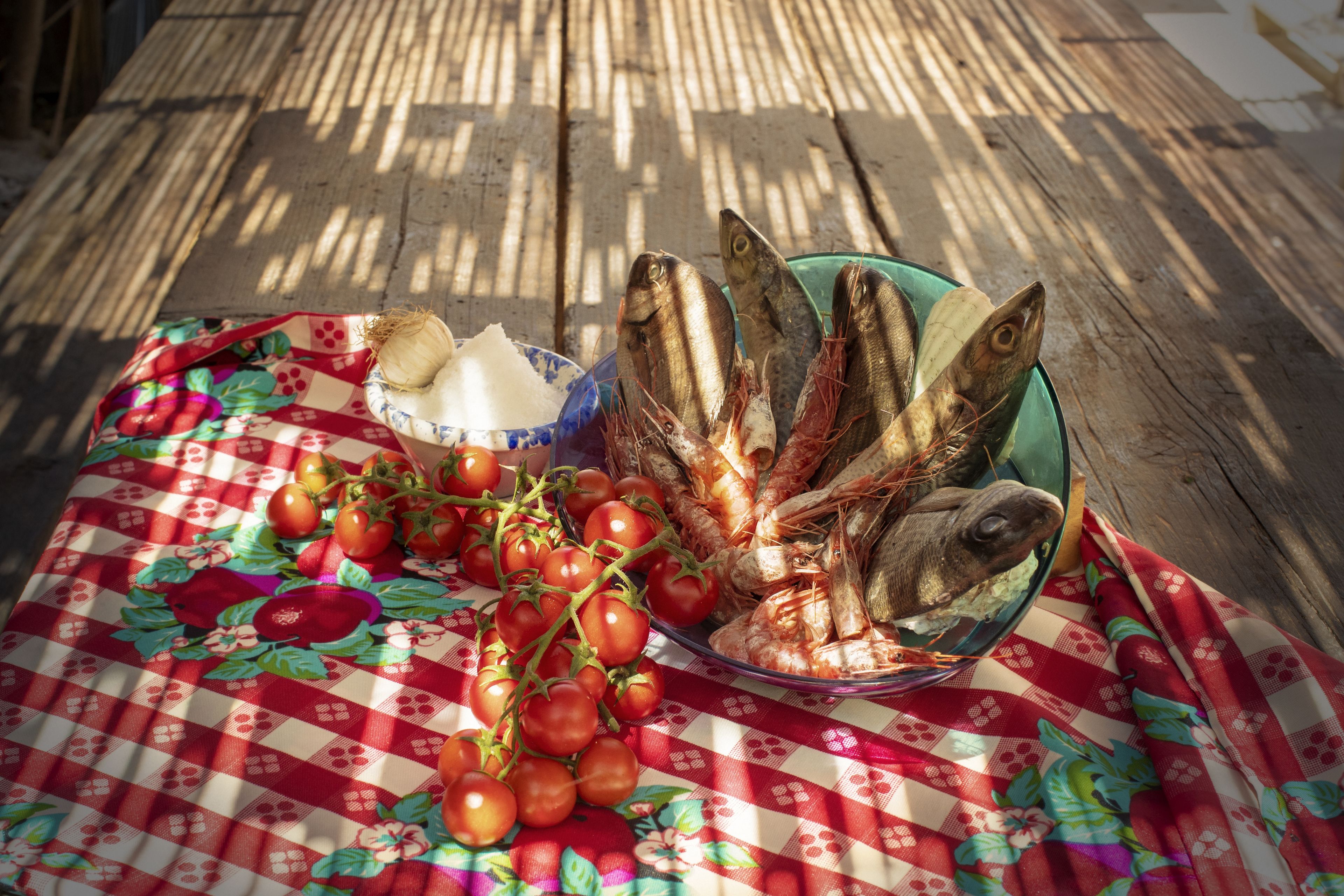 A table with a checkered tablecloth, a bowl of fish, and a bunch of tomatoes.