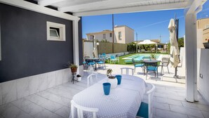 A patio with a table and chairs, a small window, and a view of a swimming pool and neighboring houses. - Migair (Misiliscemi)