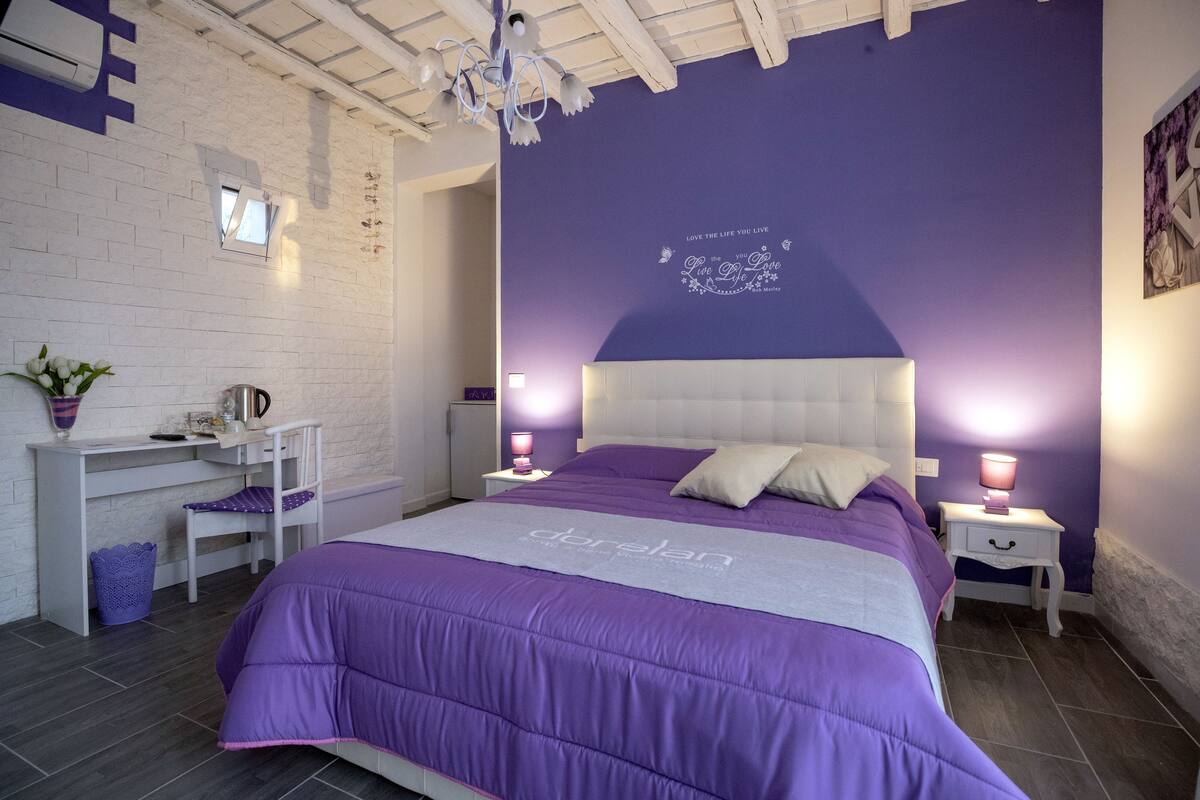 A bedroom with a purple bedspread, a white headboard, and a wall with a 'Love the life you live' quote.