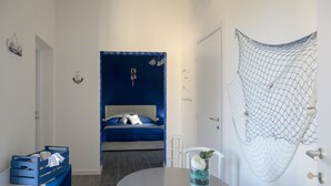 A bedroom with a bed, a bedside table with a glass of water and a flower, a blue wall, and a white door. - Migair (Misiliscemi)
