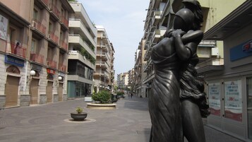A statue of a woman holding a child in a public square with multi-story buildings and shops on either side.