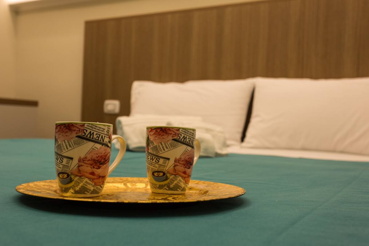 Dos tazas sobre una bandeja en una cama con colcha color turquesa y almohadas blancas.
