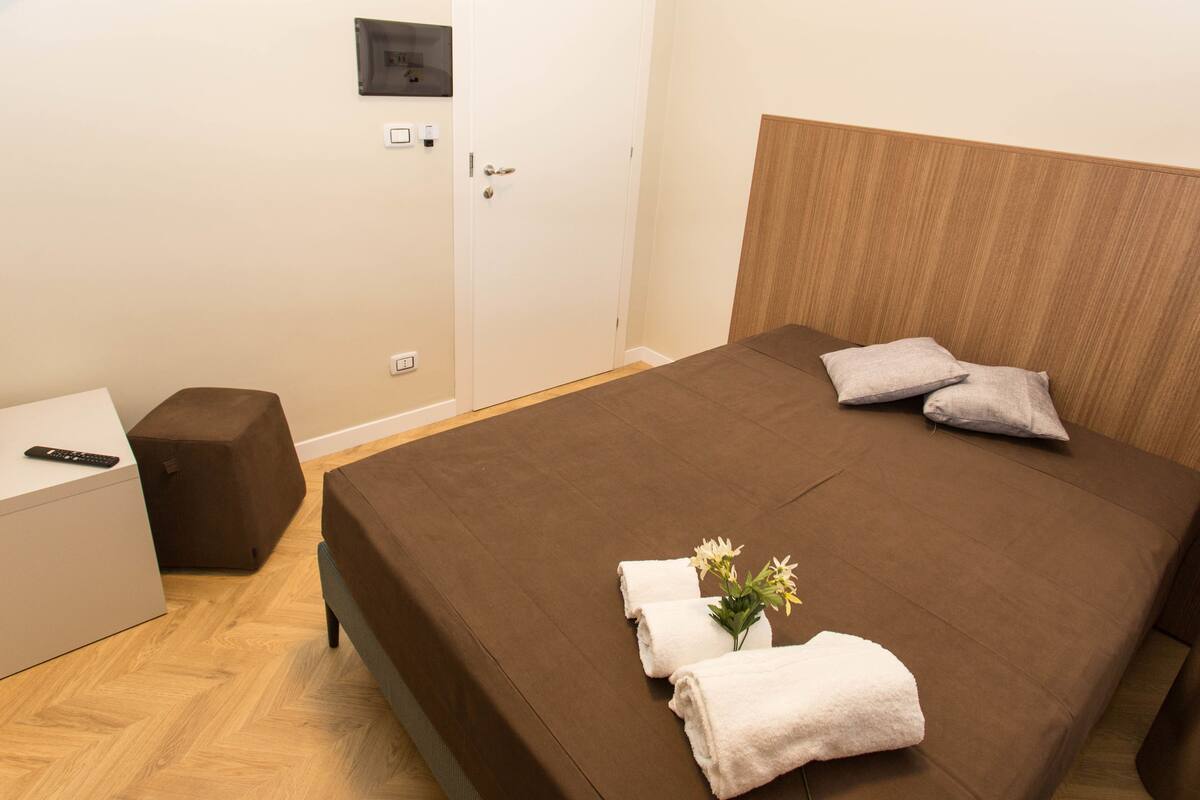 Una cama bien hecha con ropa de cama marrón, dos almohadas y un florero con flores. Hay un control remoto sobre una mesita de noche.