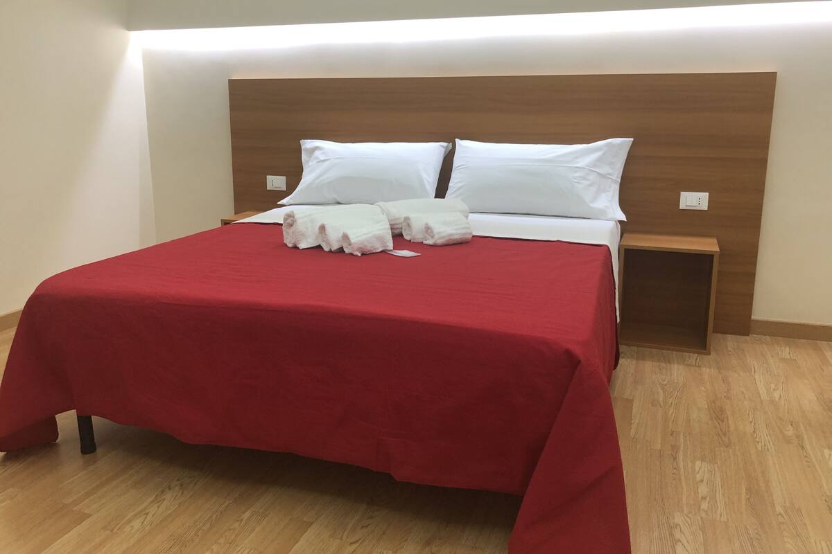 Una cama bien hecha con una colcha roja, almohadas blancas y una toalla doblada al lado.