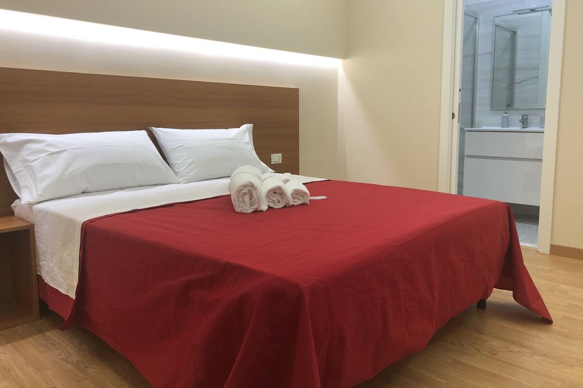 Una cama bien hecha con almohadas blancas y una colcha roja, cabecera de madera y suelo de madera.