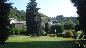 Un patio trasero con césped, una zona de sillones de jardín, un alto pino y un seto bien cuidado.