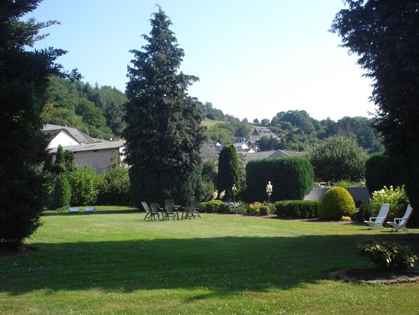 A grassy backyard with a lawn chair area, a tall coniferous tree, and a well-maintained hedge. - Eifel Hotel Lamberty (Landscheid)